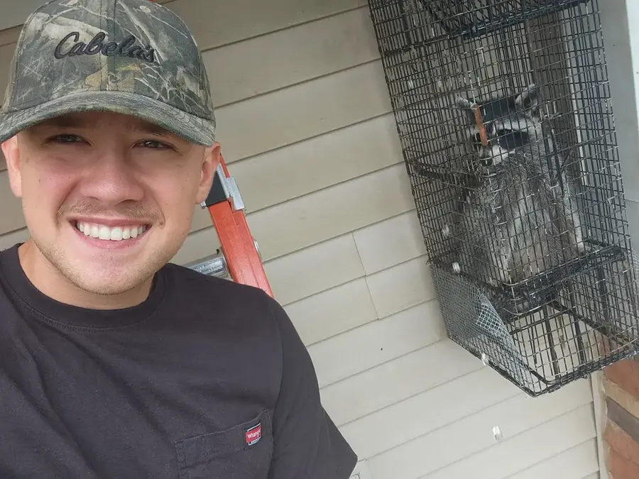 A crew member beside a raccoon in a trap against a house wall.