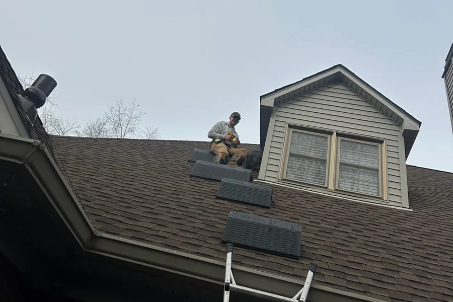 A crew member working at a dormer on a shingled roof, roof pads laid out and a ladder up against the eave.