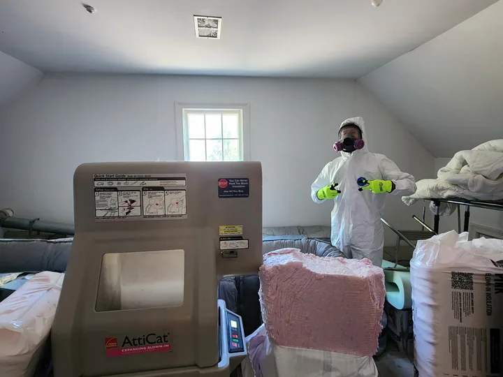 A man in a coverall and respirator working beside an insulation blowing machine in a cleared attic space.