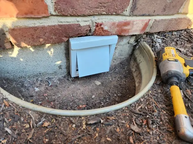 A steel dryer vent cover newly installed at the base of a brick wall.