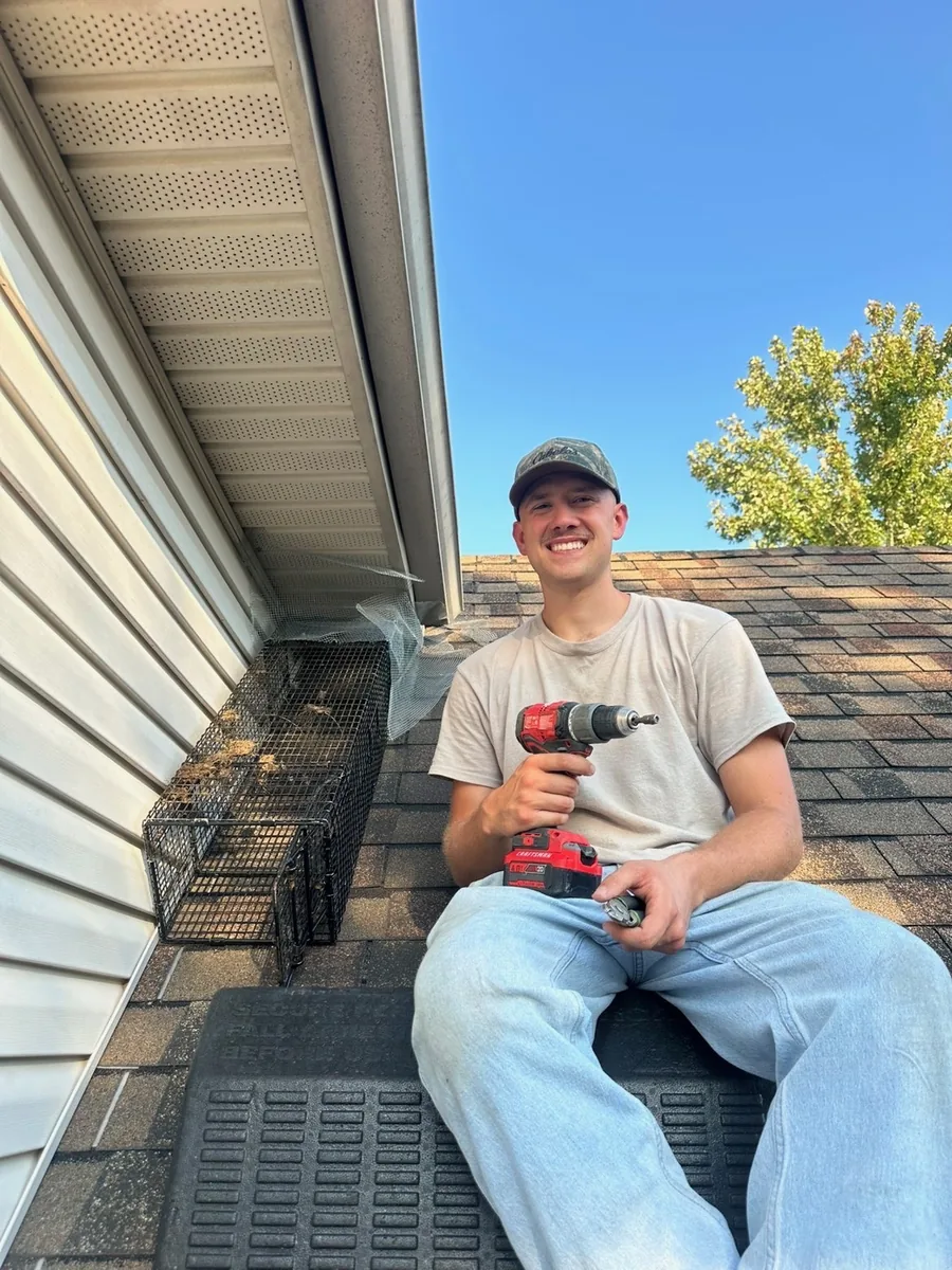 EJ sitting on a shingled roof beside a trap set against the soffit, holding a drill.